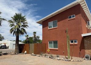 Un edificio de dos pisos con fachada roja, cerca de madera y paisaje desértico con cactus y una palmera.
