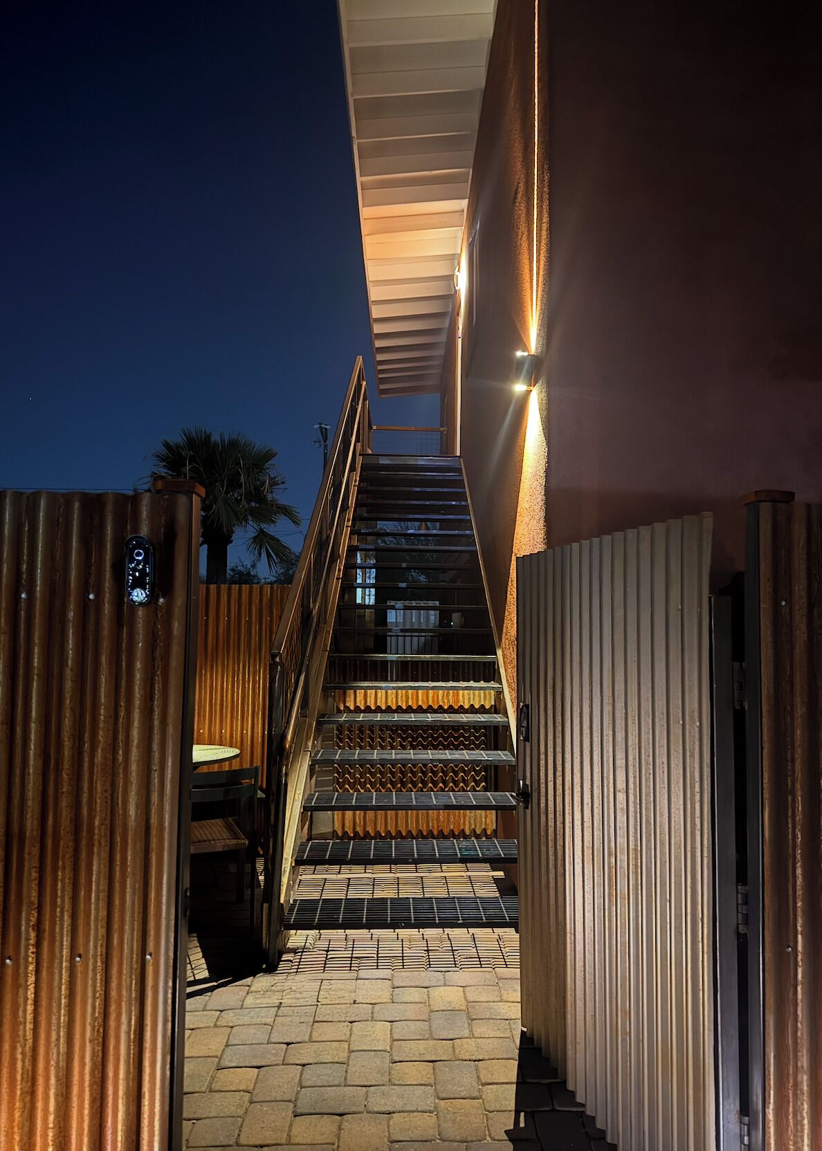 Un escalier menant à l’entrée d’un bâtiment, avec une rampe en bois et une porte en métal.