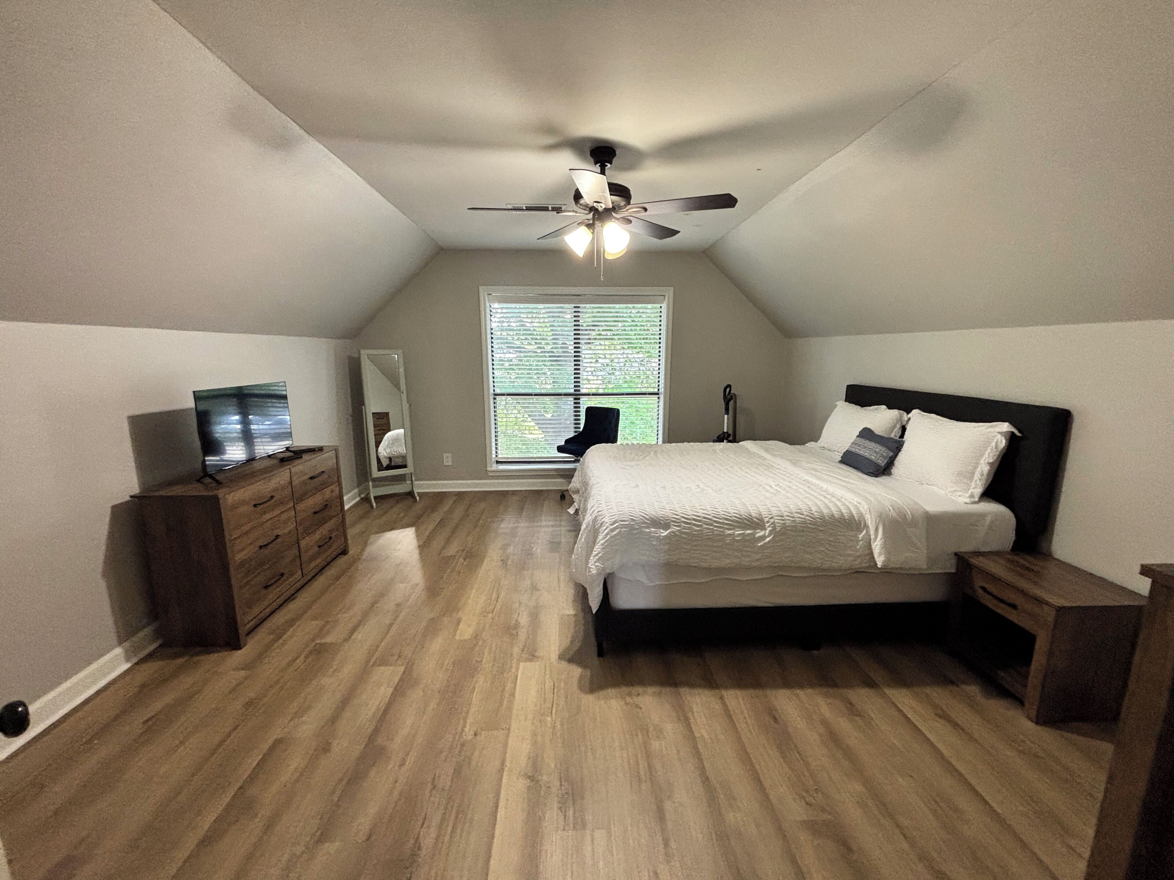 Een slaapkamer met een schuine kap, een bed met witte beddengoed, een dressoir, een spiegel en een plafondventilator.