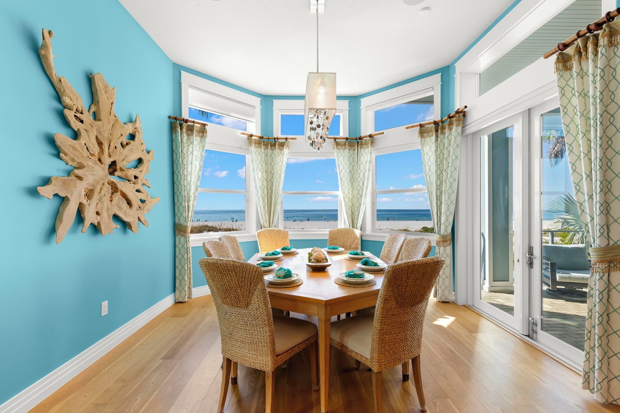 A dining area with a view of the beach, featuring a wooden table, wicker chairs, and a decorative wall piece.