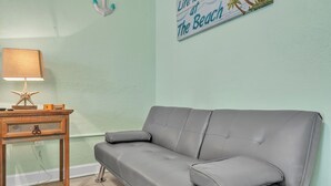 Ein Zimmer mit einem grauen Sofa, einem Holznachttisch mit einer Lampe und einer Wanddekoration mit Strandmotiv.