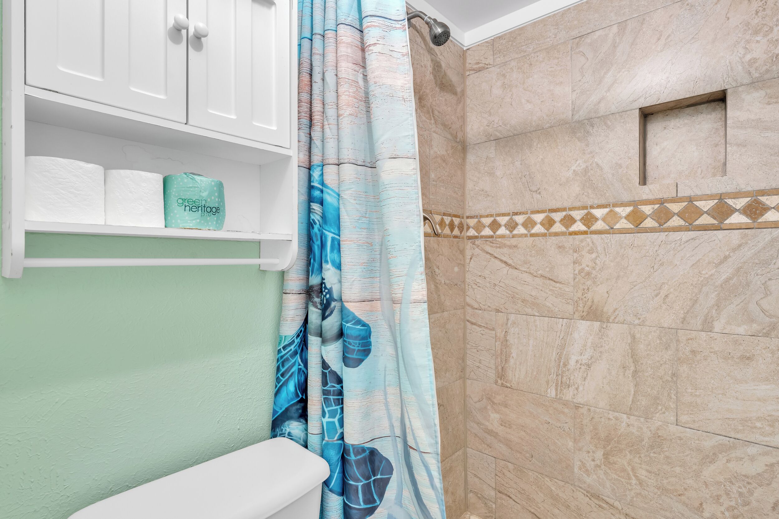 Un bagno con il water, uno scaffale con articoli da toilette, una doccia con tenda decorata e un muro piastrellato.