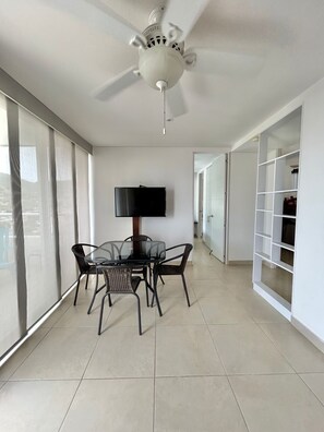 Ein geräumiges Zimmer mit Deckenventilator, einem Glastisch mit Stühlen, einem Fernseher und einem großen Fenster mit durchsichtigen Vorhängen.
