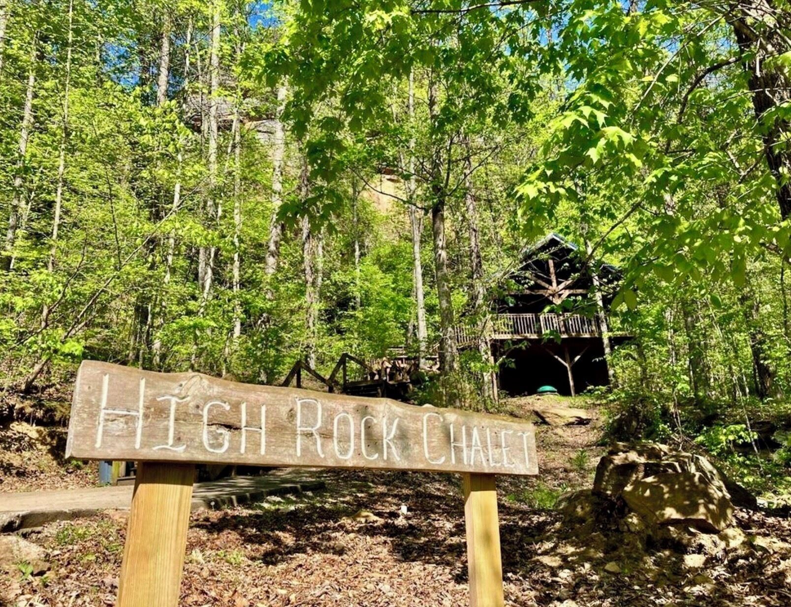 A wooden sign with 'HIGH ROCK CHALET' written on it, surrounded by a forest with a cabin in the background.