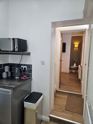 A compact kitchen with a microwave, kettle, and coffee maker. A trash bin is visible, and a small table with a plant is seen in the adjacent room. - Nancys Nook - Whitby (Whitby)