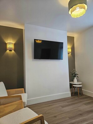 A modern living room with a flat-screen TV mounted on the wall, two wall-mounted lamps, a round side table with a vase, and a wicker sofa. - Nancys Nook - Whitby (Whitby)