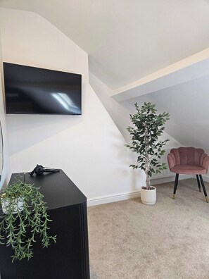 A modern living room with a flat-screen TV mounted on the wall, a potted plant, a black side table, a pink chair, and a carpeted floor. - Salt & Sage - Whitby (Whitby)