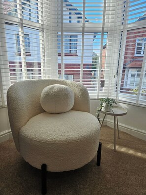 A room with a large window, a round chair with a cushion, a small table with a plant, and a carpeted floor. - Salt & Sage - Whitby (Whitby)