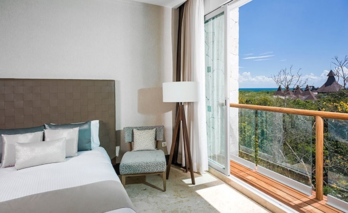 A hotel room with a bed, a chair, a lamp, and a balcony with a view of the sea and buildings.