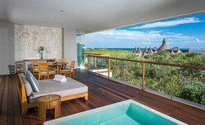 Un balcone con pavimento in legno, una zona relax con divano e poltrone, un piccolo tavolino con un vaso e vista sull'oceano e su edifici con tetti di paglia.