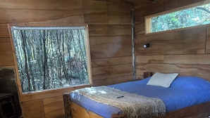 Ein Interieur einer Holzhütte mit einem Bett, einem Fenster mit Blick auf einen Wald, einem roten Sessel und einem Holztisch.