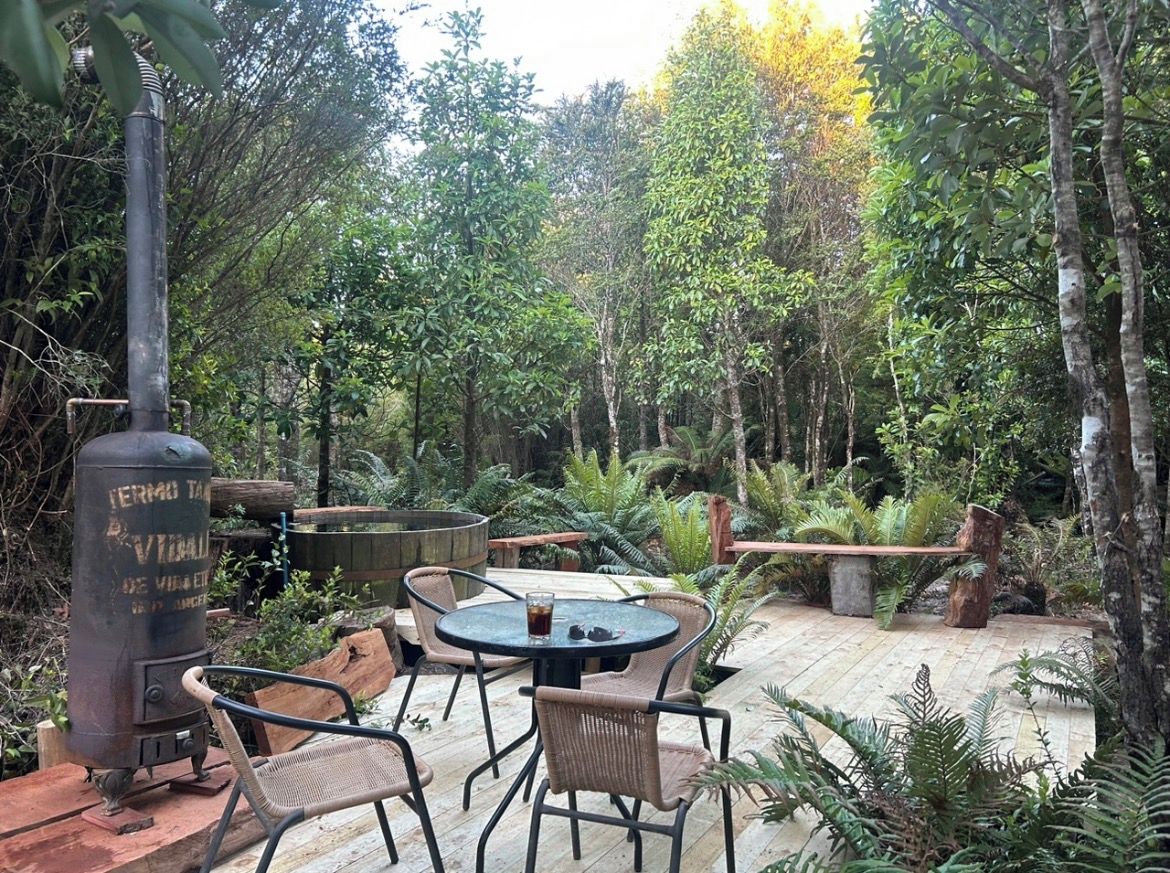 Een terras met een ronde tafel en stoelen, omgeven door weelderige begroeiing en bomen.
