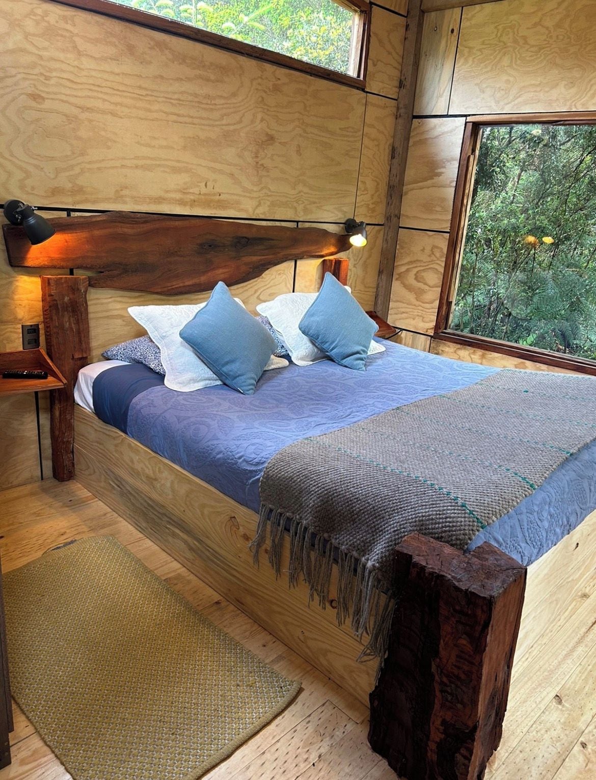 A wooden room with a bed, a nightstand, a window, and a view of greenery.