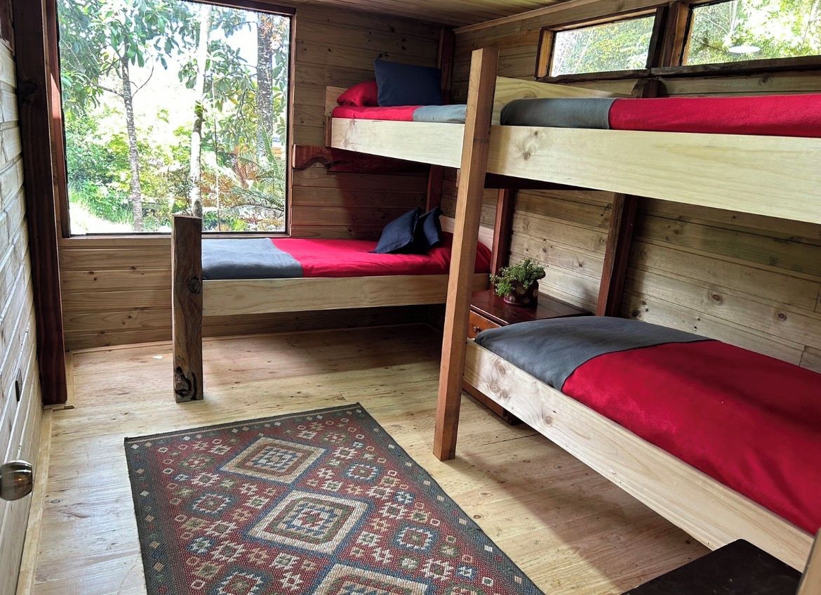 Interior de cabana com beliches, piso de madeira, tapete estampado e janela com vista para árvores.
