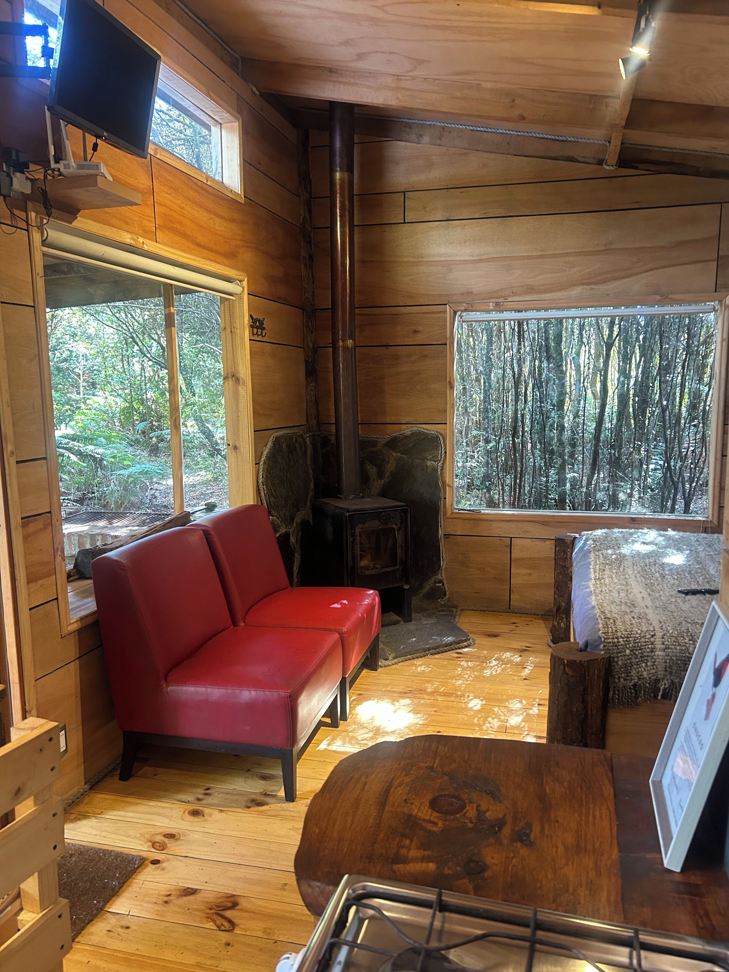 Interior de una cabaña con un sofá rojo, una estufa de leña y un ventanal con vistas al bosque.