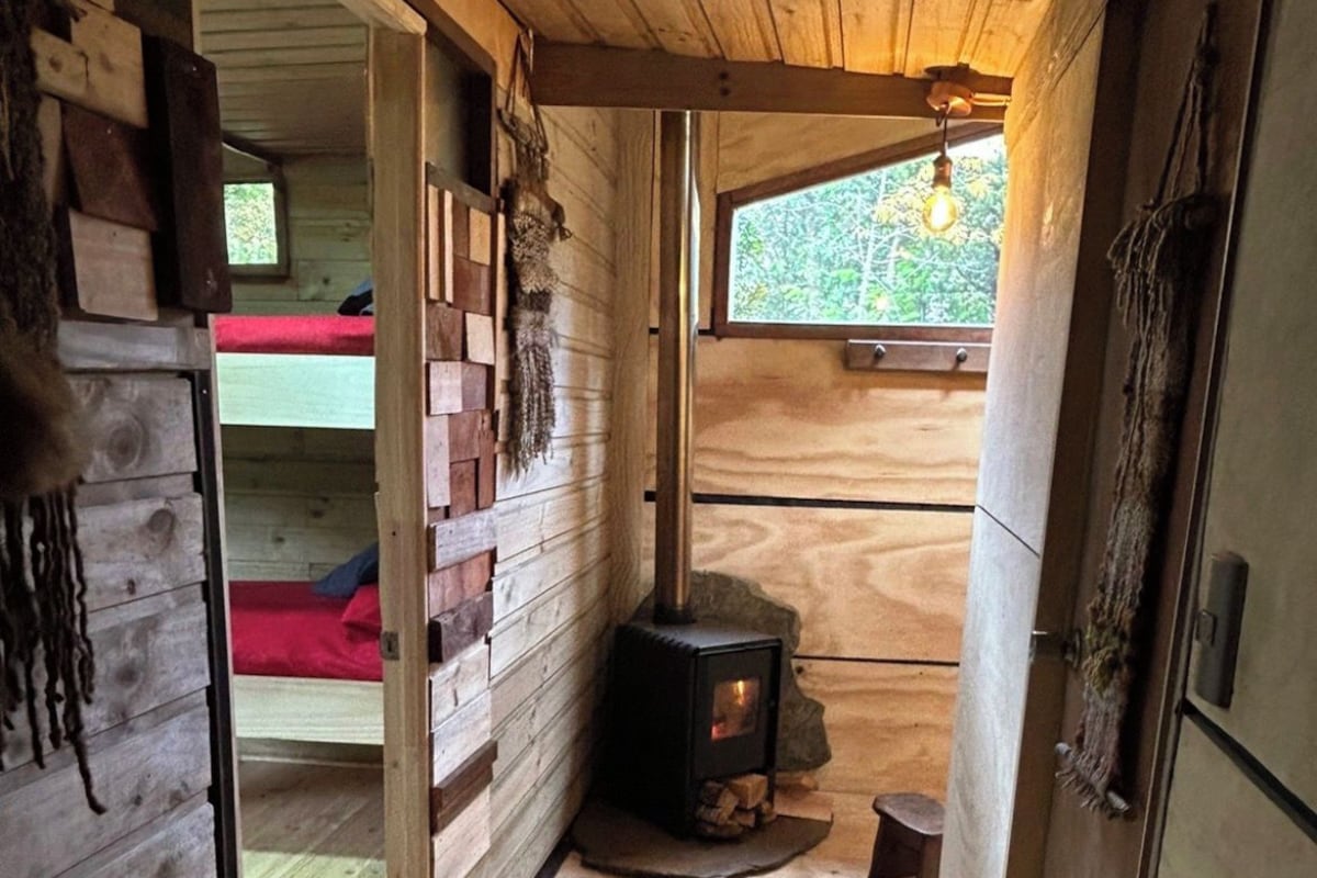 Un interior de madera con chimenea, una cama roja y un ventanal con vistas a la vegetación.