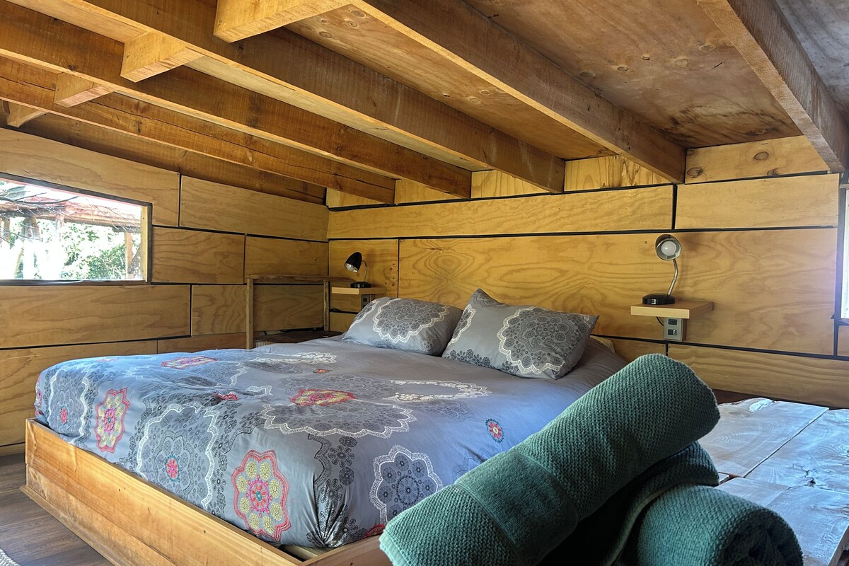 Un dormitorio de madera con cama, almohadas, mesita de noche y ventana.