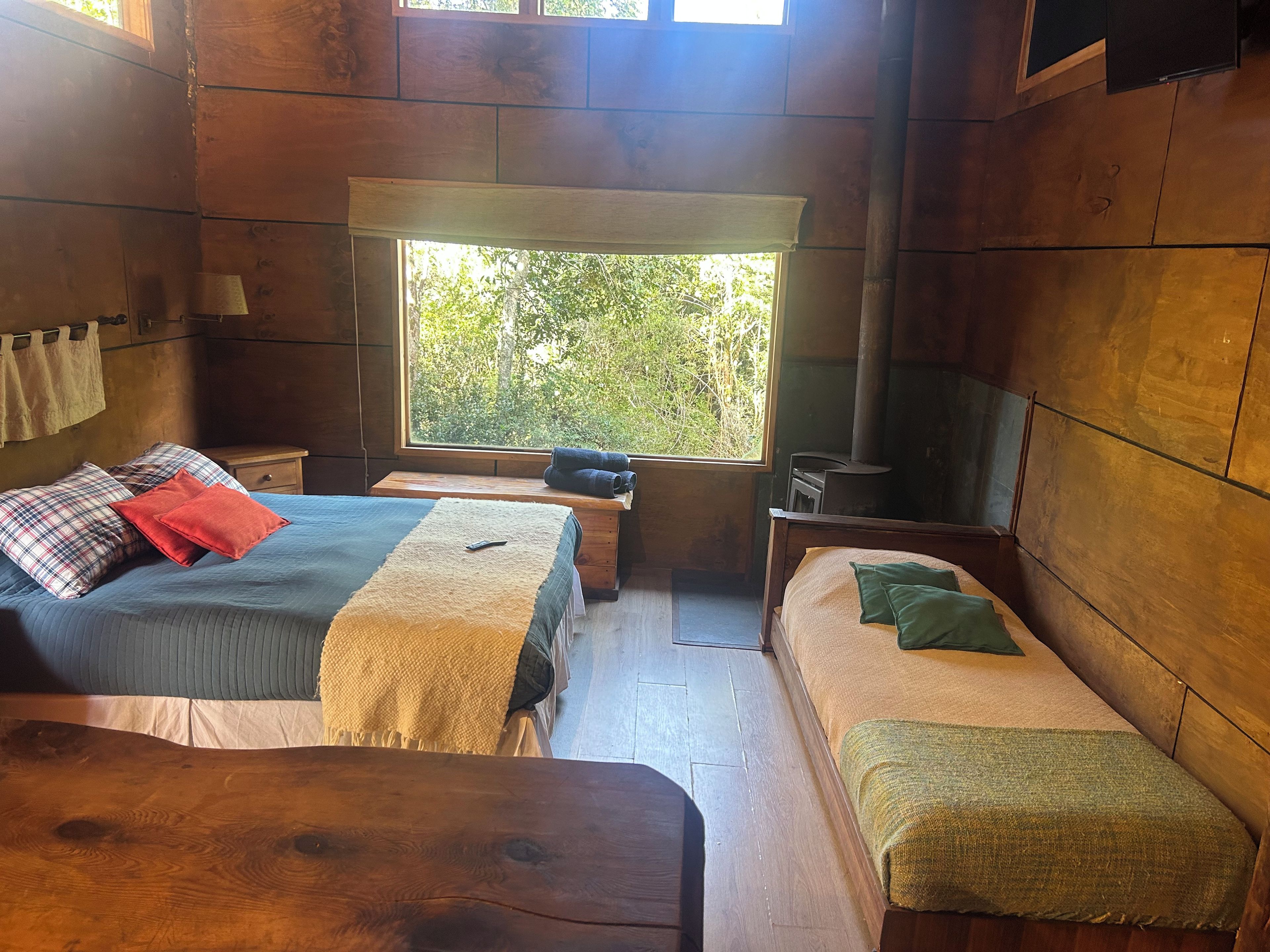 A cozy cabin room with two beds, a wooden bench, a small table, a window with a view of greenery, and a wood-burning stove.
