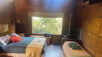 A cozy cabin room with two beds, a wooden bench, a small table, a window with a view of greenery, and a wood-burning stove.