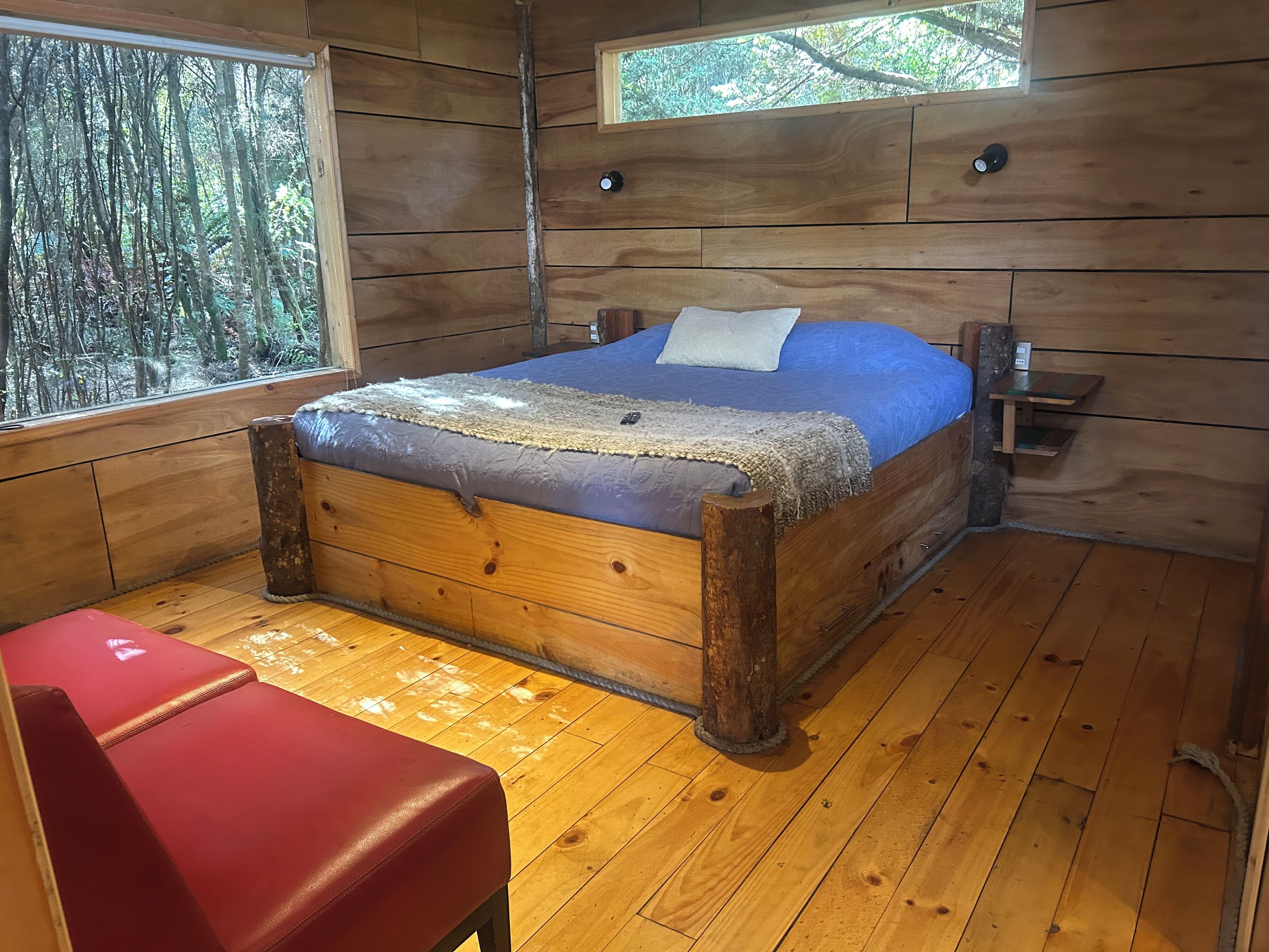 Un dormitori de cabana de fusta amb un llit, una banca vermella i una finestra amb vista a un bosc.