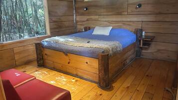 Un dormitori de cabana de fusta amb un llit, una banca vermella i una finestra amb vista a un bosc.