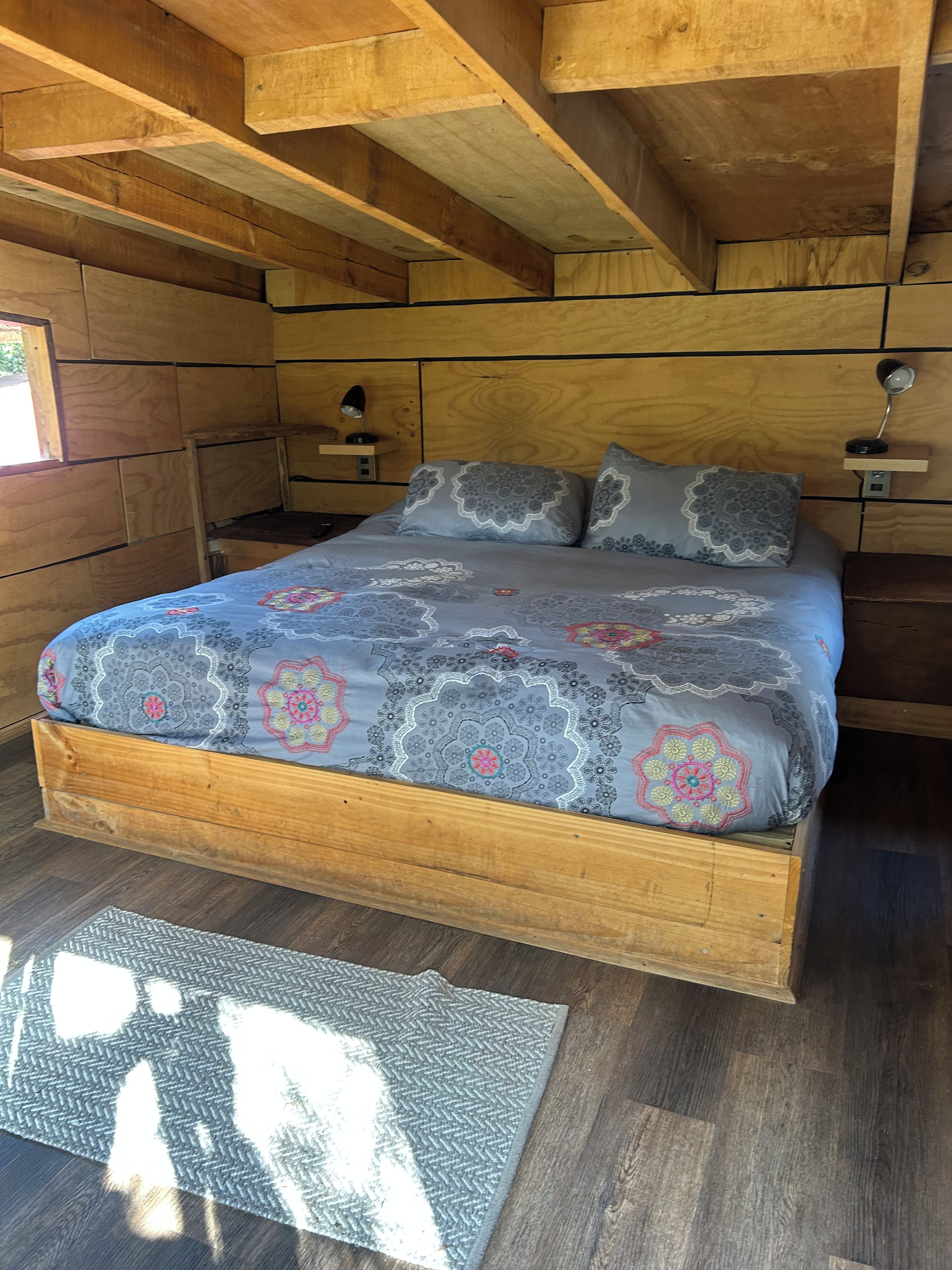A bedroom with a wooden bed frame, a patterned bedspread, a bedside table with a lamp, and a nightstand.