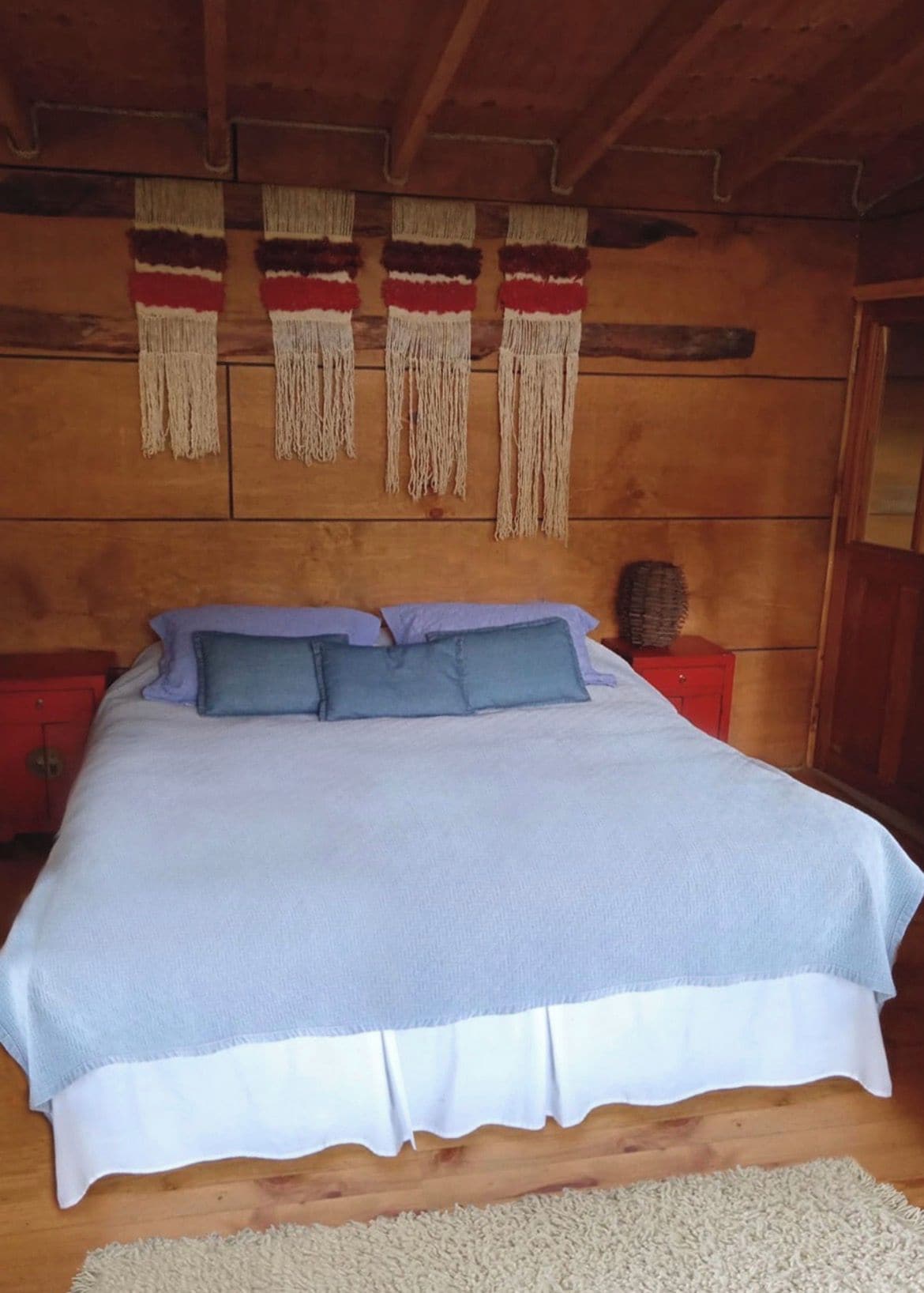 A wooden-framed bedroom with a bed, pillows, a nightstand, and macramé wall hangings.