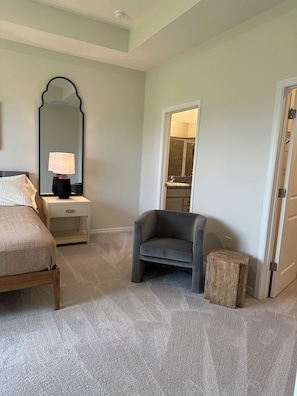 Une chambre à coucher comprenant un lit, une table de chevet, une lampe, une chaise et une porte donnant sur une salle de bain.