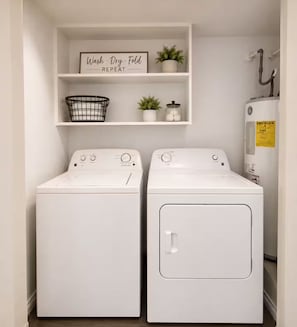 A laundry room with two white front-loading washers and dryers, a shelf with a 'Wash, Dry, Fold, REPEAT' sign, a black wire basket, and a small potted plant.