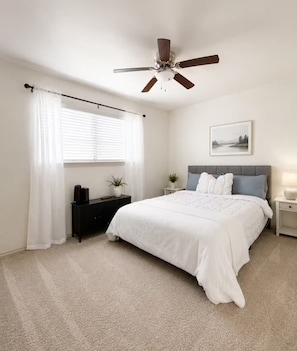 A bedroom with a bed, a ceiling fan, a window with curtains, a bedside table, and a nightstand.