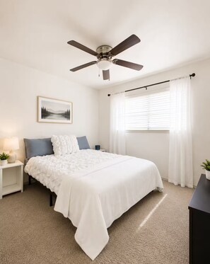 Un dormitorio con cama, mesita de noche, lámpara, ventilador de techo, ventana con cortinas y un cuadro en la pared.