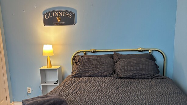 Uma cama com um edredom acolchoado escuro, uma luminária dourada em uma mesa de cabeceira e um anúncio da Guinness na parede.