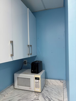 A compact kitchen with a microwave on a marble countertop, a toaster on top of it, and a blue wall with white cabinets.
