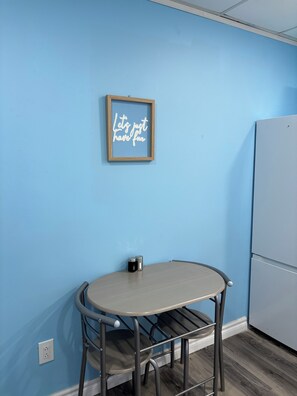 A small room with a blue wall, a framed inspirational quote, a round table with two chairs, a small black container, and a white refrigerator.