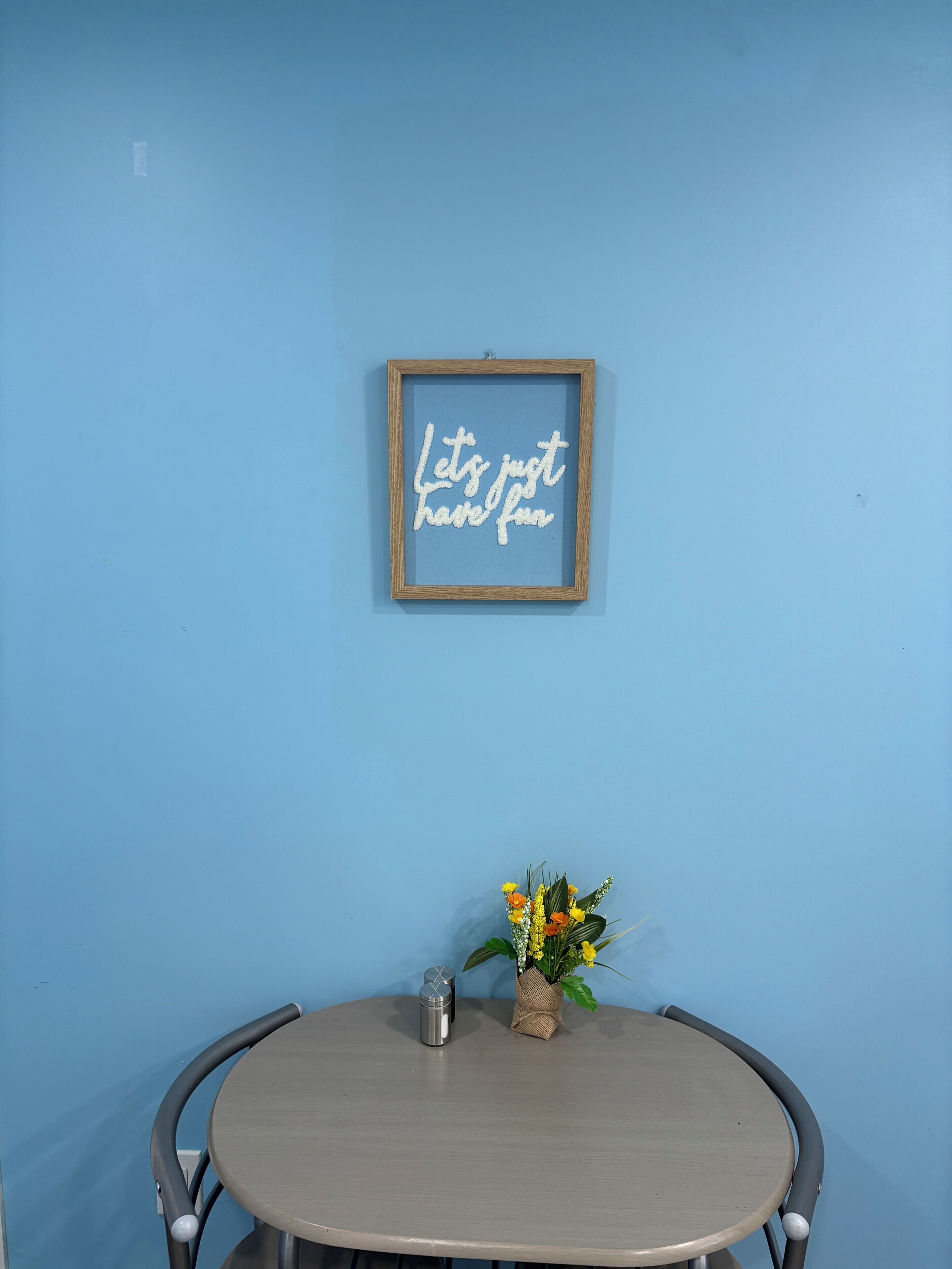 A round table with a metal chair, a framed sign on the wall, and a small plant with flowers.