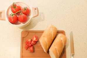 A wooden cutting board with two baguettes and sliced tomatoes, a knife, and a bowl of tomatoes on a speckled countertop. - Spacious 3BR Family Home | Bunks + Ping Pong (Peoria)