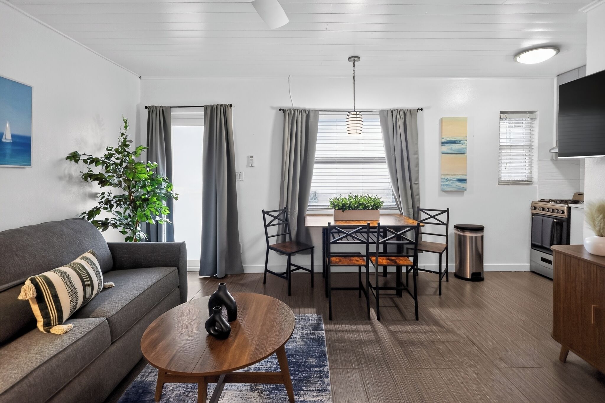 Een moderne woonkamer met een bank, salontafel, eettafel en stoelen. Er staat een plant, een schilderij en er is een keuken met een fornuis en kasten.