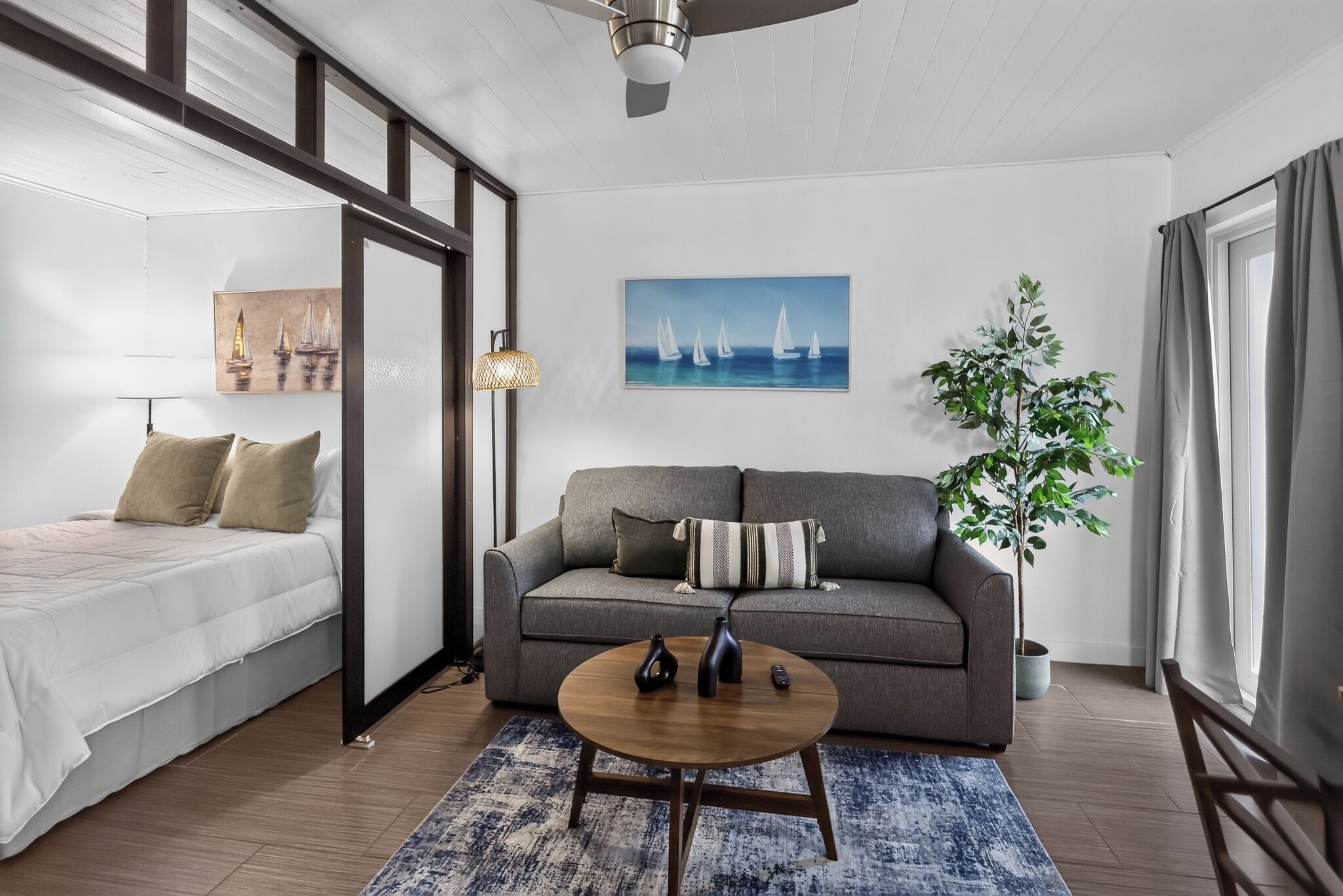 A modern room with a bed, a sofa, a coffee table, and a painting of sailboats.