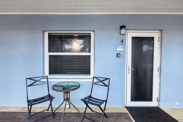 Pared exterior azul con una puerta blanca y una ventana con persianas. Hay una pequeña mesa y dos sillas frente a la ventana.