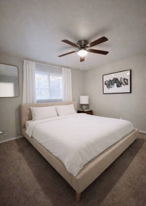 A bedroom with a bed, a ceiling fan, a window with curtains, a bedside table with a lamp, and a framed picture on the wall. - Comfortable 2BR Retreat - 1st Floor Living (Waxahachie)