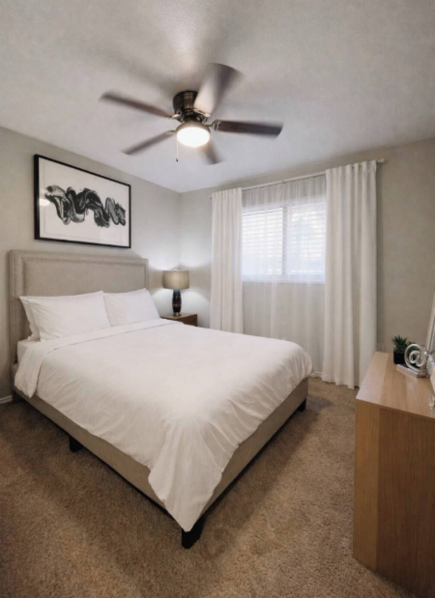 Une chambre d’hôtel avec un lit, un ventilateur de plafond, une fenêtre avec des rideaux, une lampe de chevet et un tableau encadré au mur.