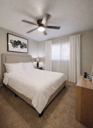 A hotel room with a bed, a ceiling fan, a window with curtains, a bedside lamp, and a framed picture on the wall. - Comfortable 2BR Retreat - 1st Floor Living (Waxahachie)