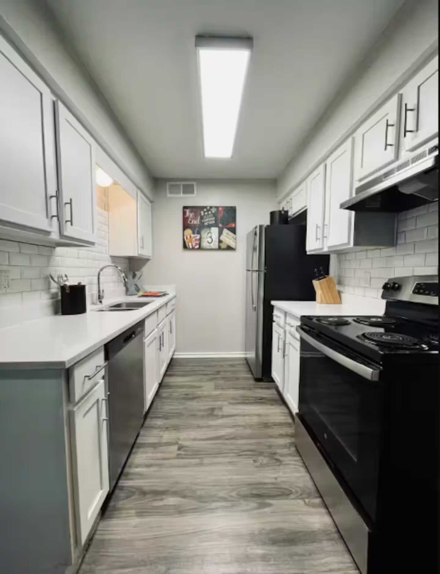 A narrow kitchen with white cabinets, a black refrigerator, and a wooden countertop.