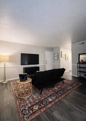A living room with a black sofa, a flat-screen TV mounted on the wall, a patterned rug, and a small round side table. - Comfortable 2BR Retreat - 1st Floor Living (Waxahachie)