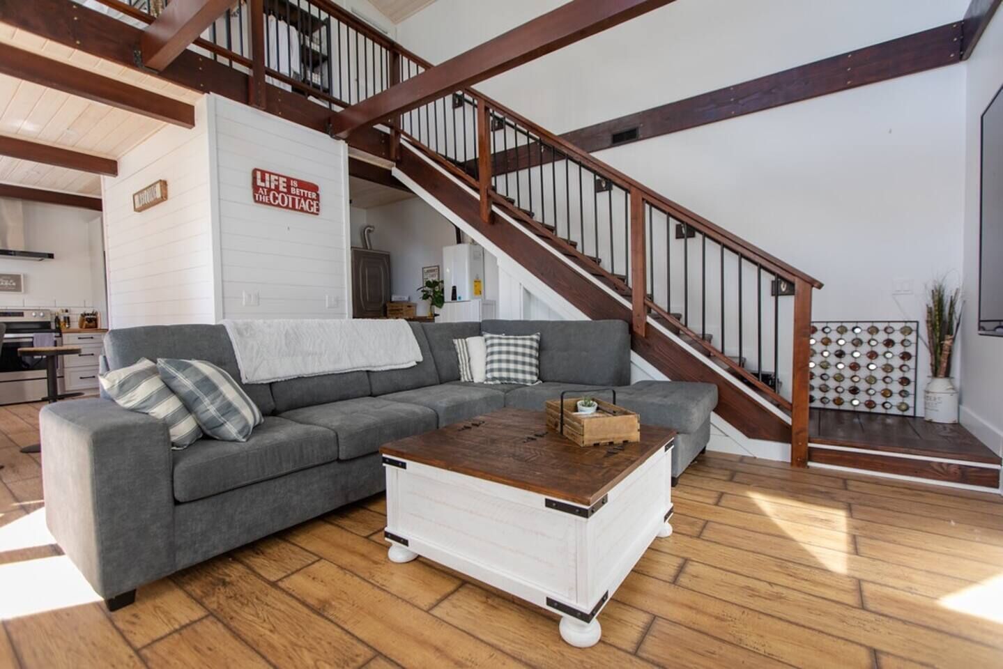 Una sala moderna con un sofá seccional gris, un aparador blanco y una mesa de centro de madera. Una escalera de madera con barandilla de metal conduce a un nivel superior.