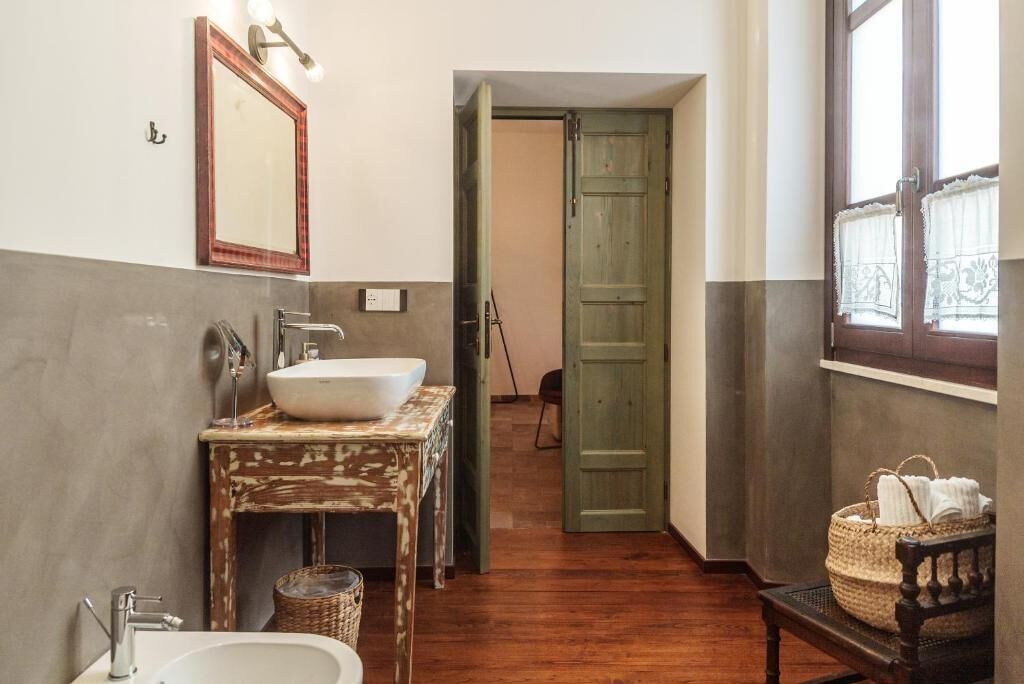 A bathroom with a large white sink, a mirror with lights, a wooden door, and a wicker basket on a bench.