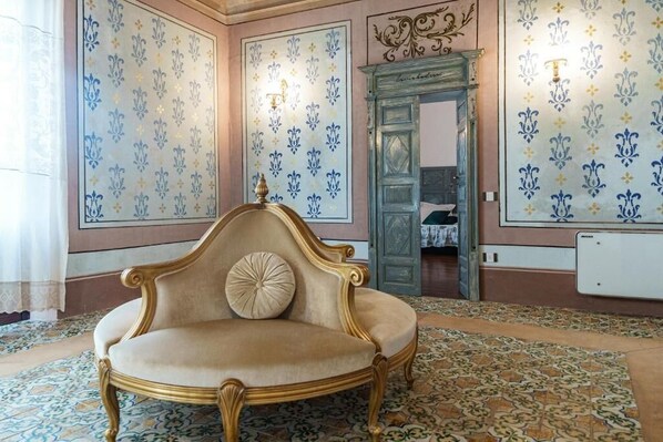 A room with a classic chaise lounge, patterned wallpaper, and a traditional fireplace.
