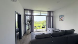 Ein modernes Wohnzimmer mit einem grauen Sofa, einer Topfpflanze und Blick auf eine grüne Landschaft durch große Fenster.