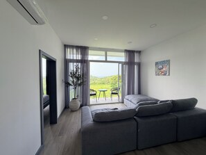 A modern living room with a grey sofa, a potted plant, and a view of a green landscape through large windows. - Retreat with views of the mountains and the sea - Roatán (Jonesville)