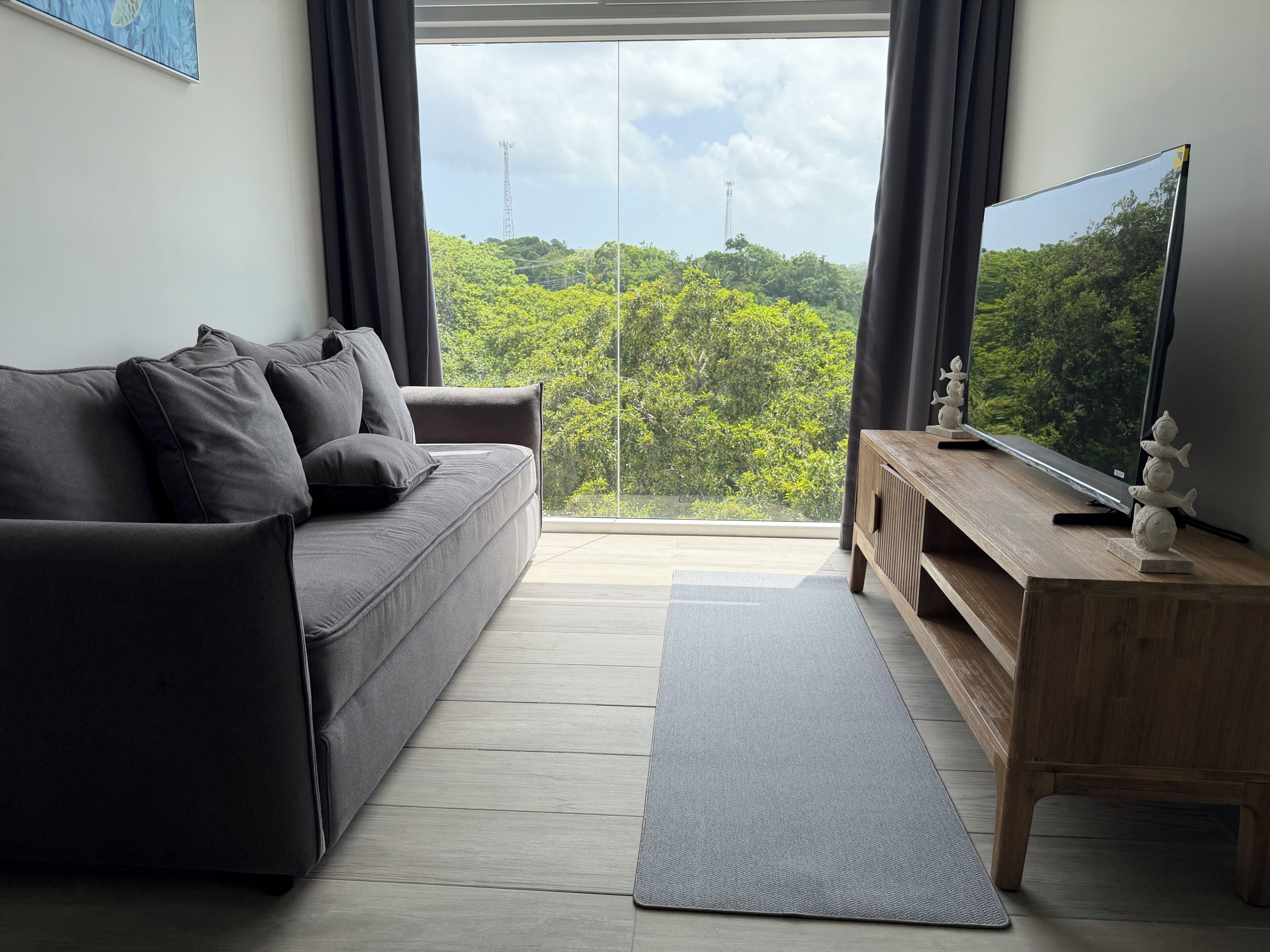 Una sala de estar moderna con un sofá gris, un mueble de televisión de madera y vistas a la vegetación a través de un gran ventanal.